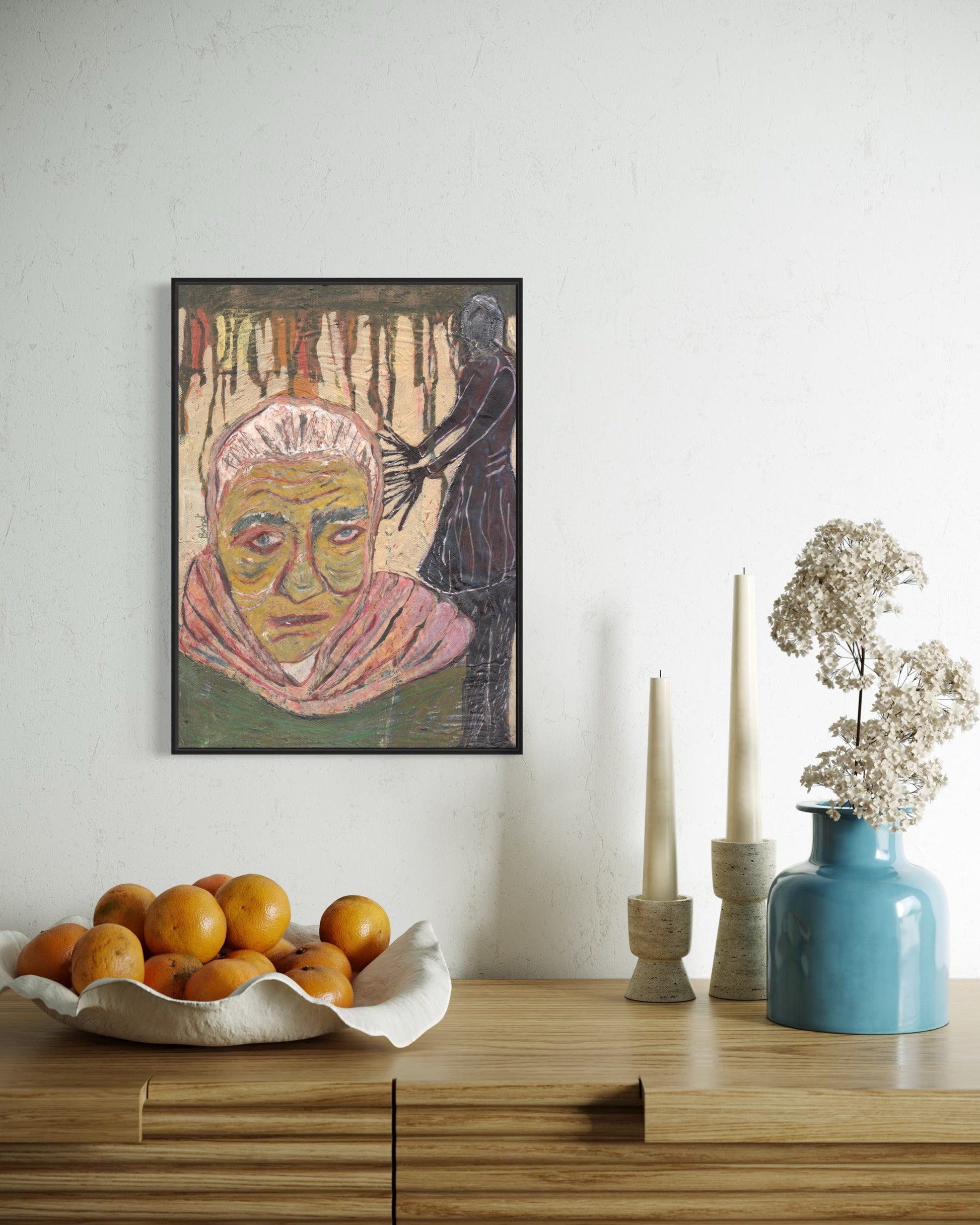 Framed artwork on a wall above a wooden surface with a bowl of fruit, candles, and a vase.