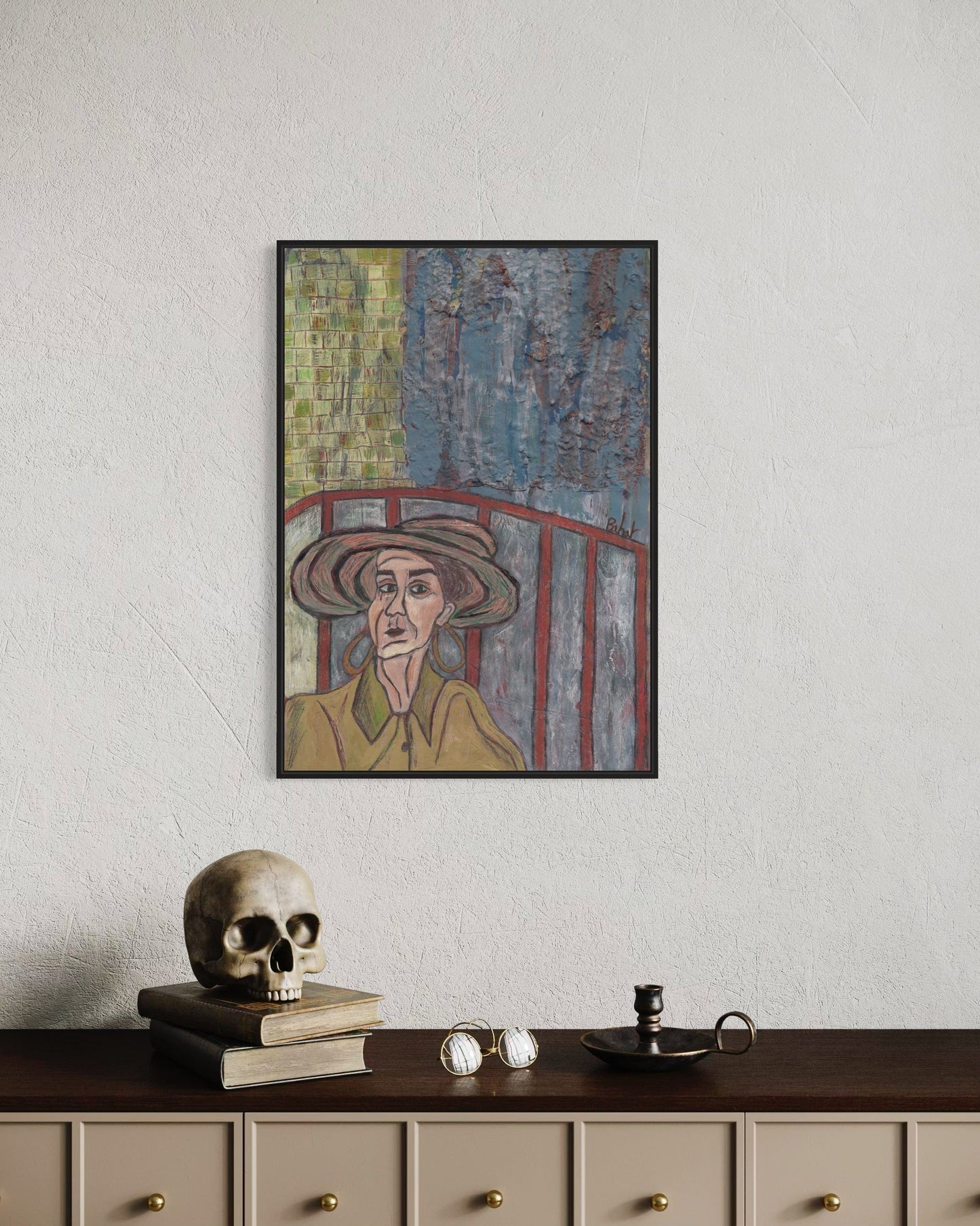 Framed artwork of a person wearing a hat on a wall above a desk with a skull and books.