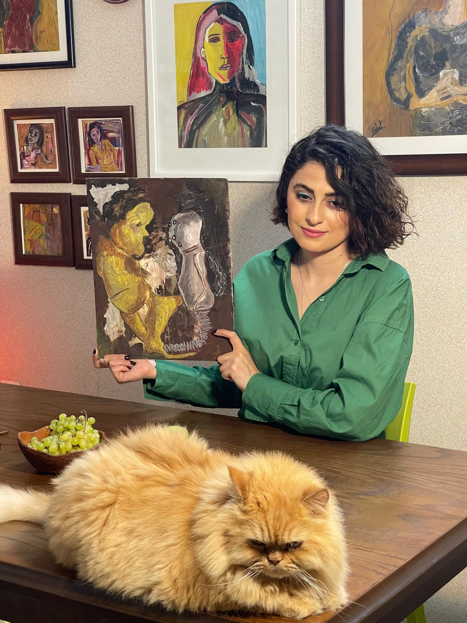 Woman in green shirt pointing at a painting with a fluffy cat on a table in front of her.