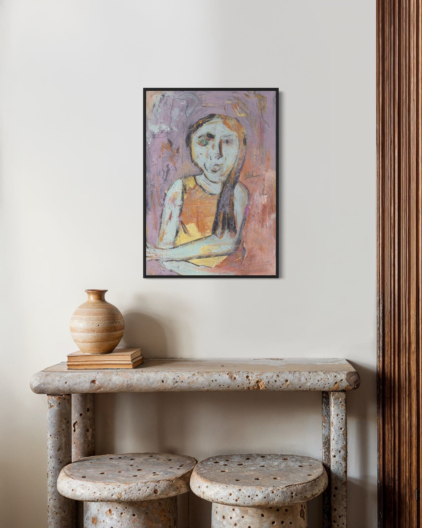 Framed abstract painting on a wall above a rustic table with a vase and books.