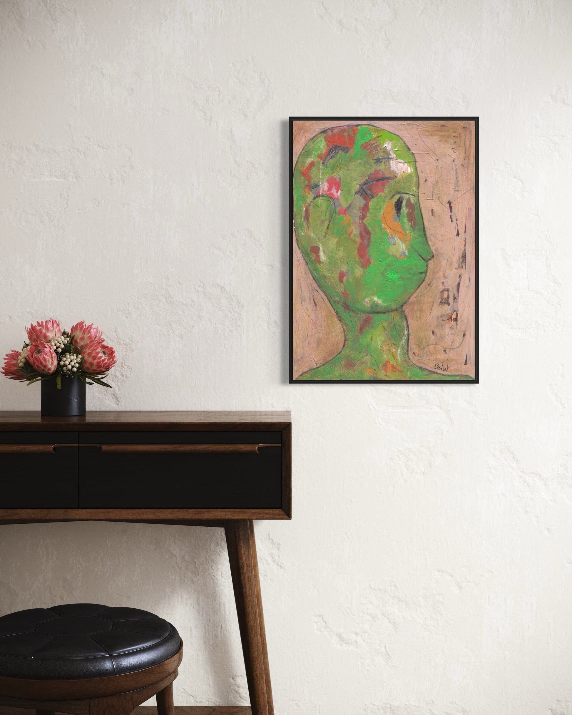 Framed artwork of a stylized face on a wall above a wooden desk with a black stool.