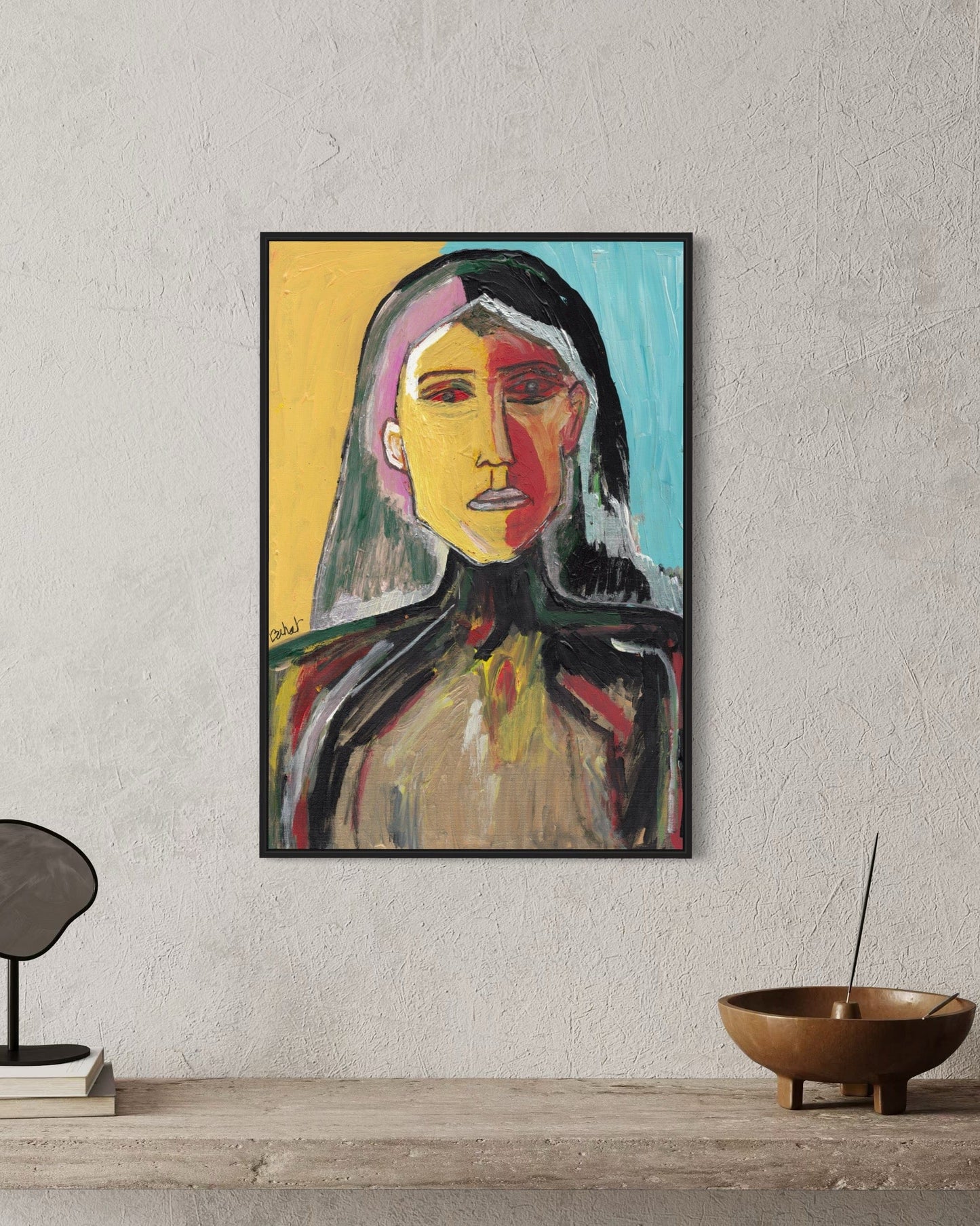 Framed abstract painting of a woman's portrait on a wall with a bowl and books on a shelf below.