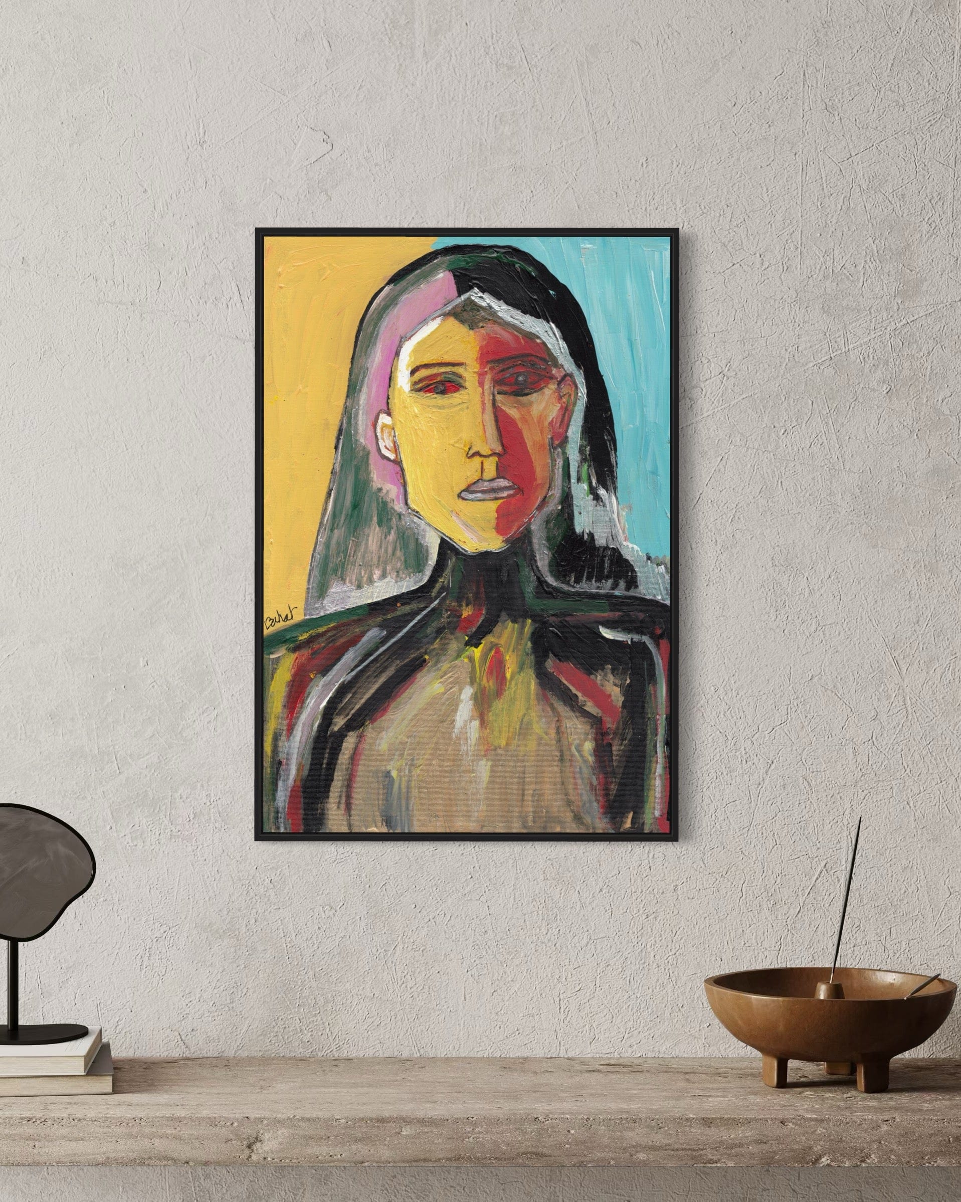 Framed abstract painting of a woman's portrait on a wall with a bowl and books on a shelf below.