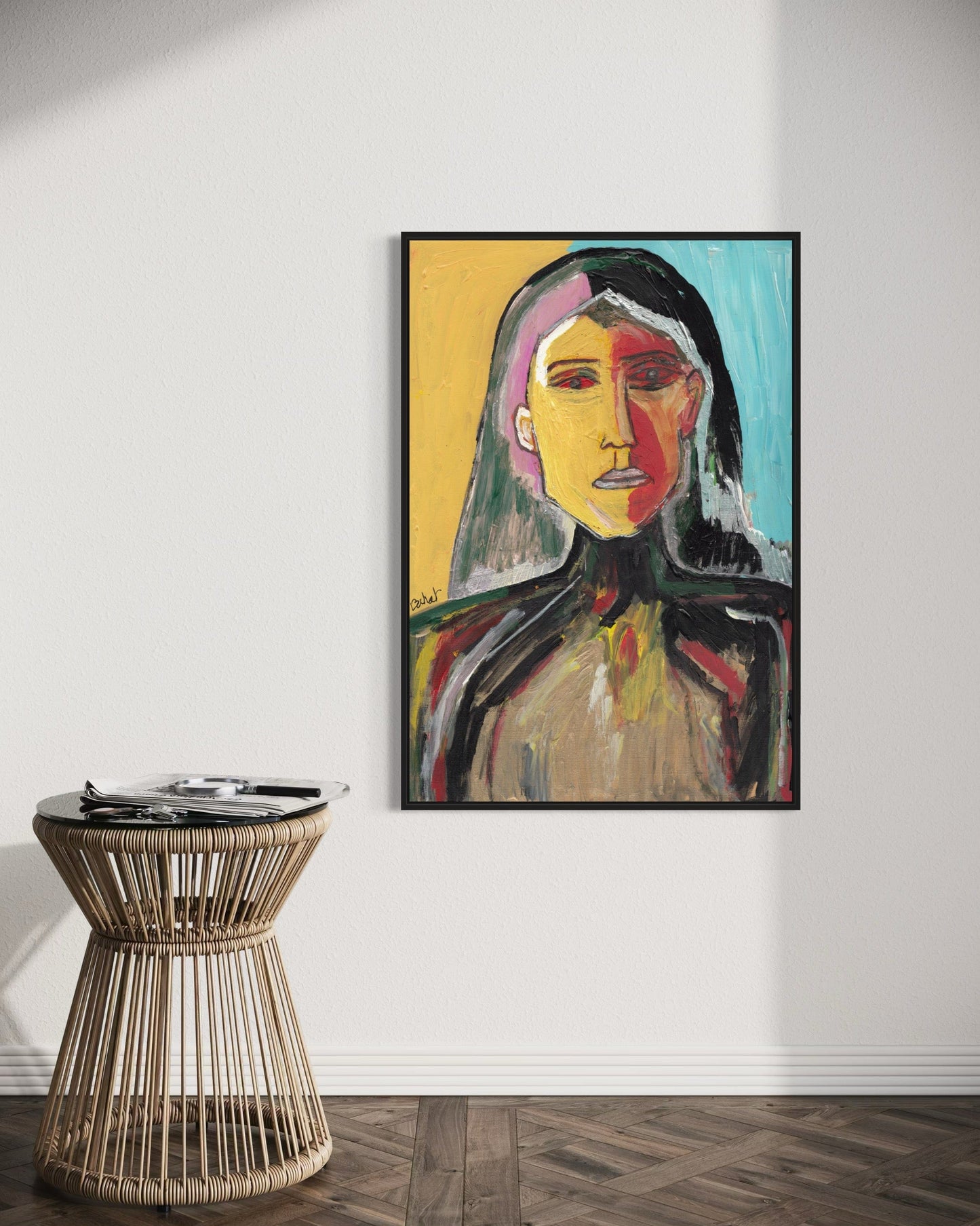 Framed abstract art of a face on a white wall with a wicker side table below.