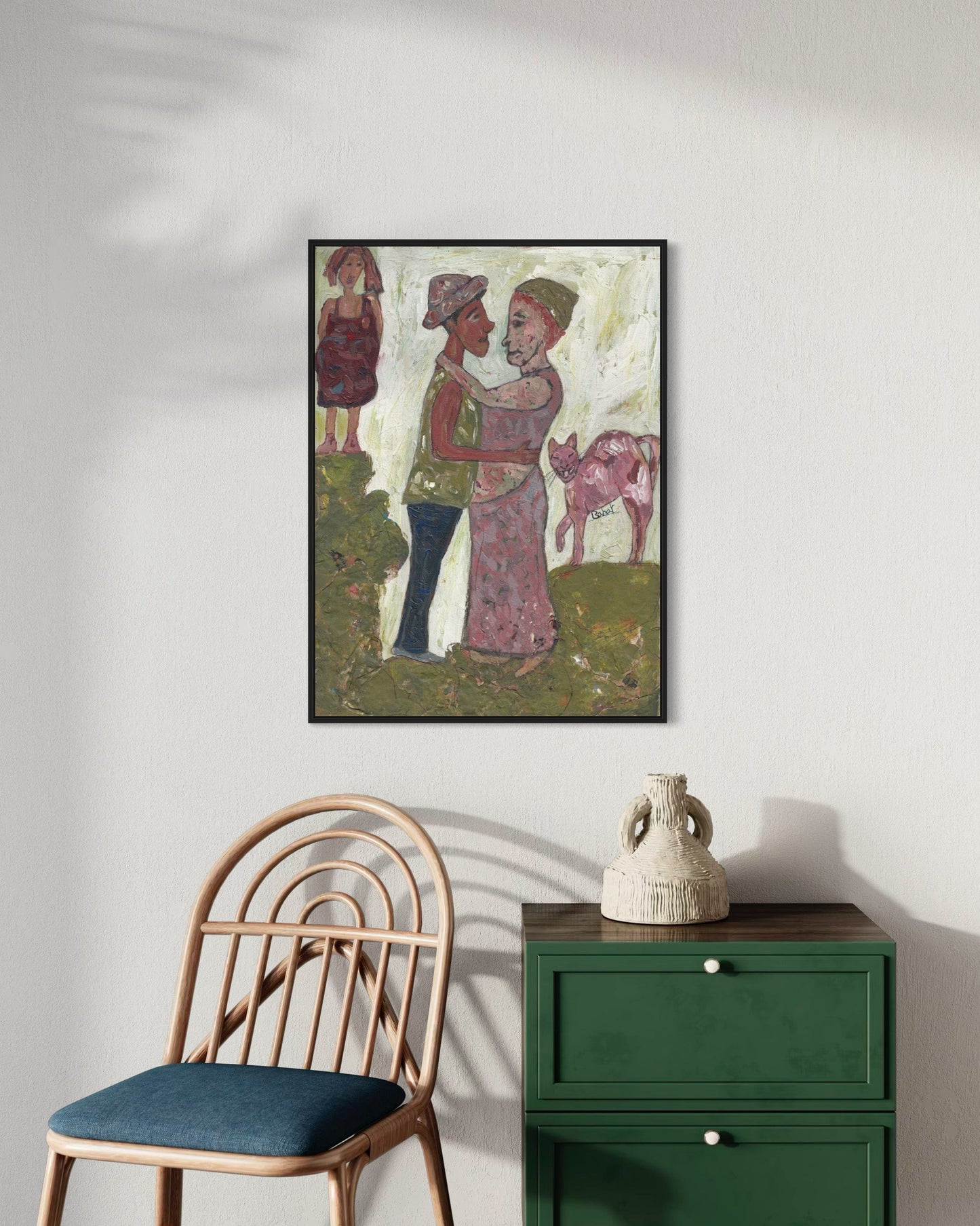 Framed artwork of two people embracing with a child and cow, placed above a green cabinet and wooden chair.