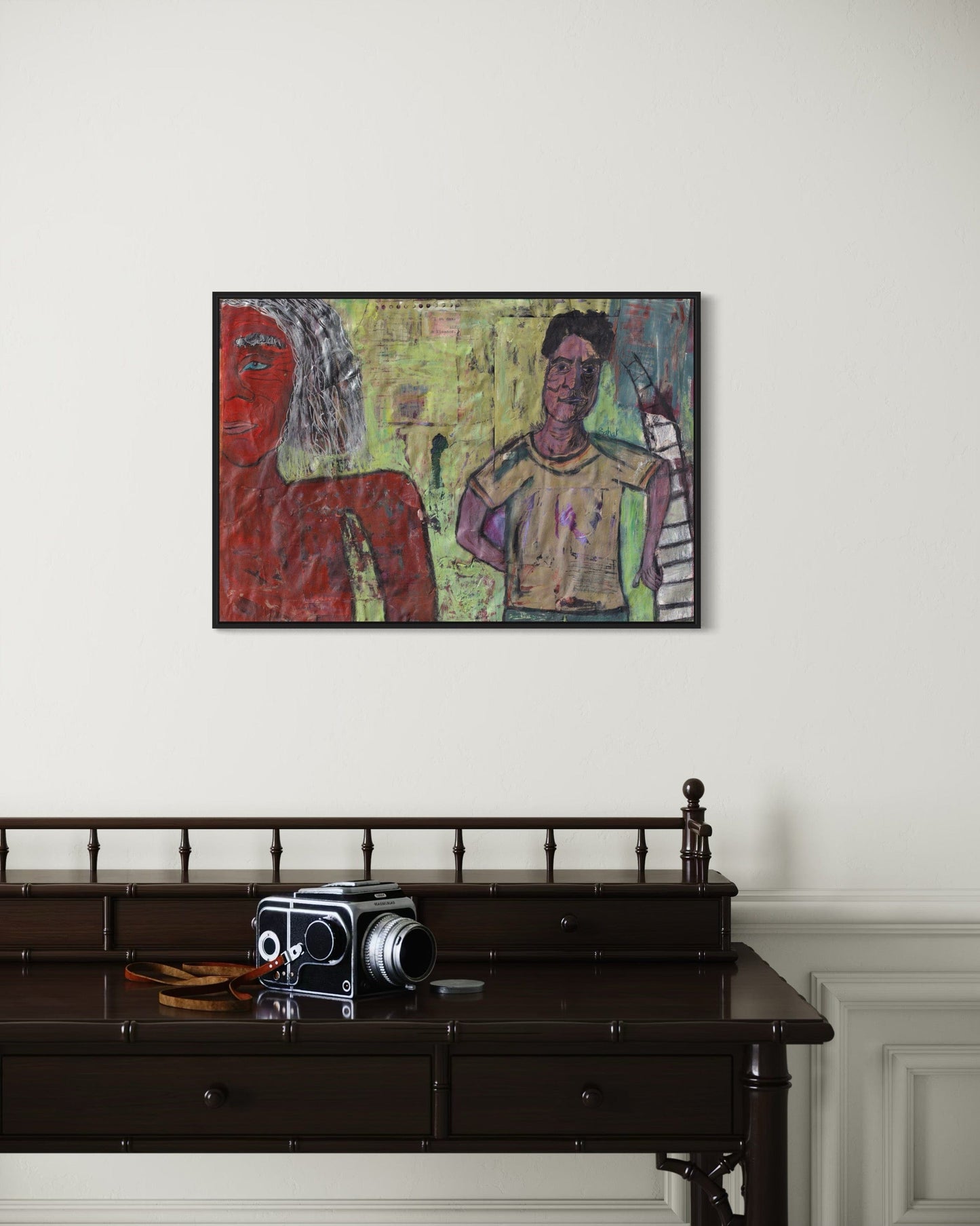 Framed artwork of two figures on a wall above a dark wooden desk with an old camera.