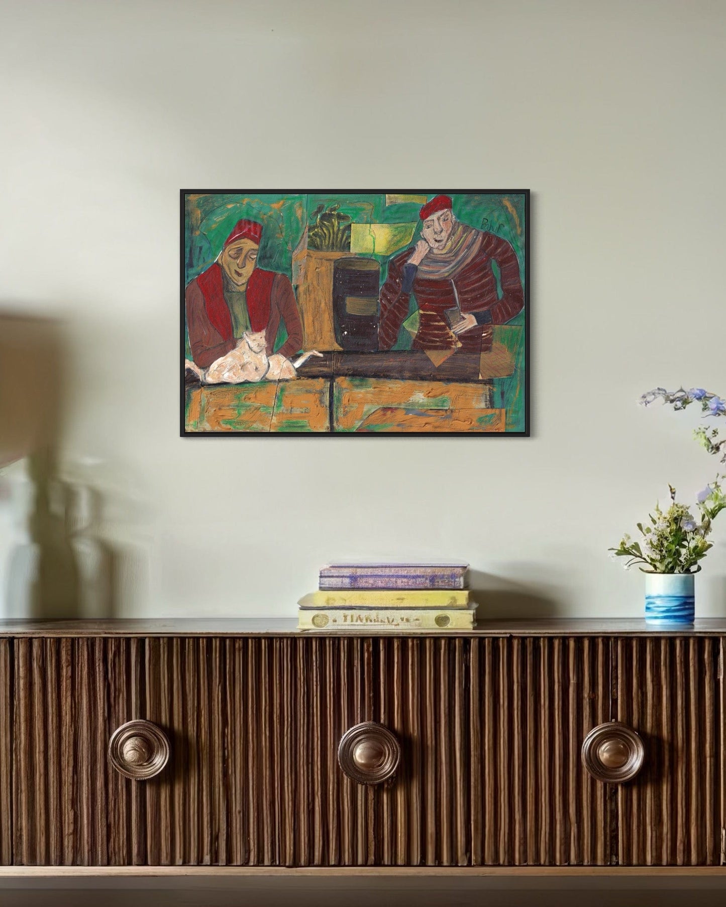 Framed artwork on a wall above a wooden cabinet with books and a plant.