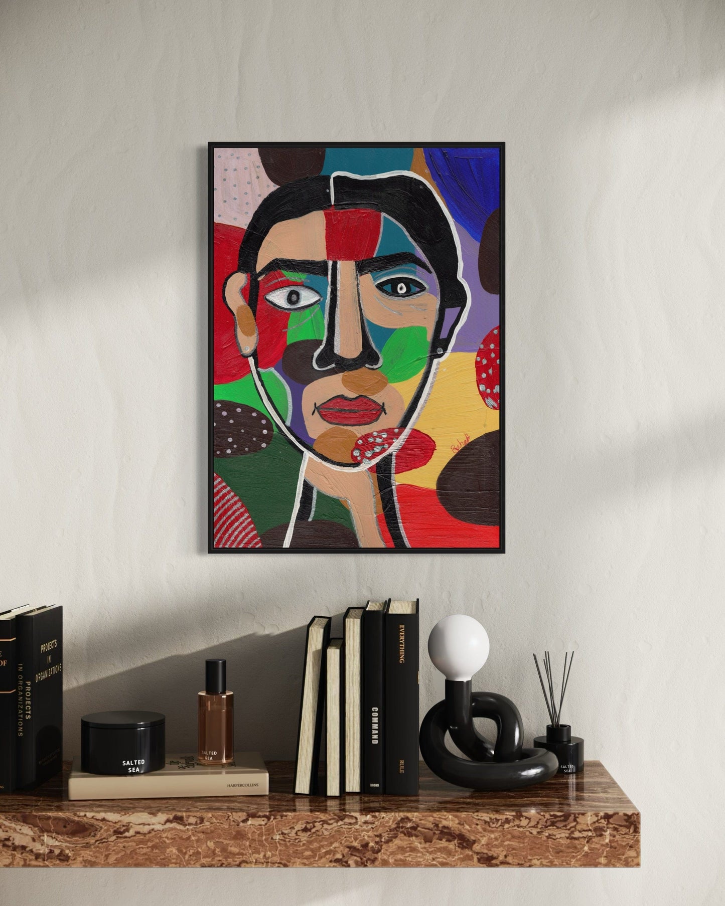Colorful abstract art print of a face on a wall above a shelf with books and decor items.