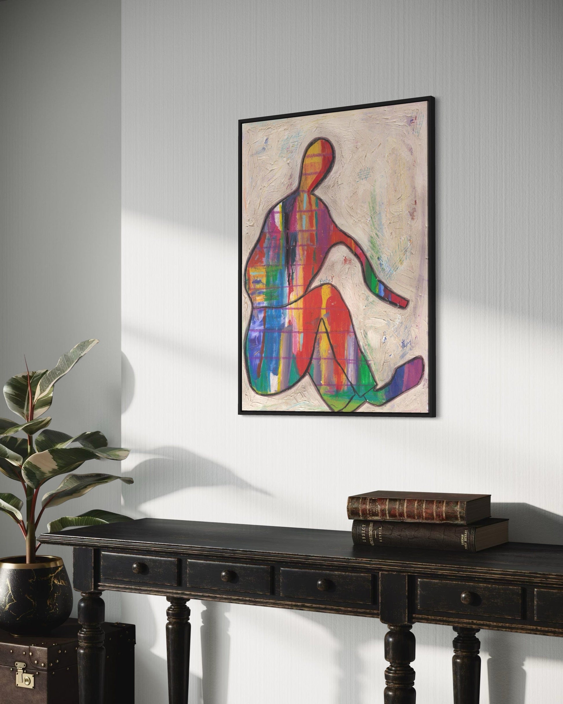Colorful abstract artwork of a sitting figure on a wall above a dark wooden table with books and a plant.