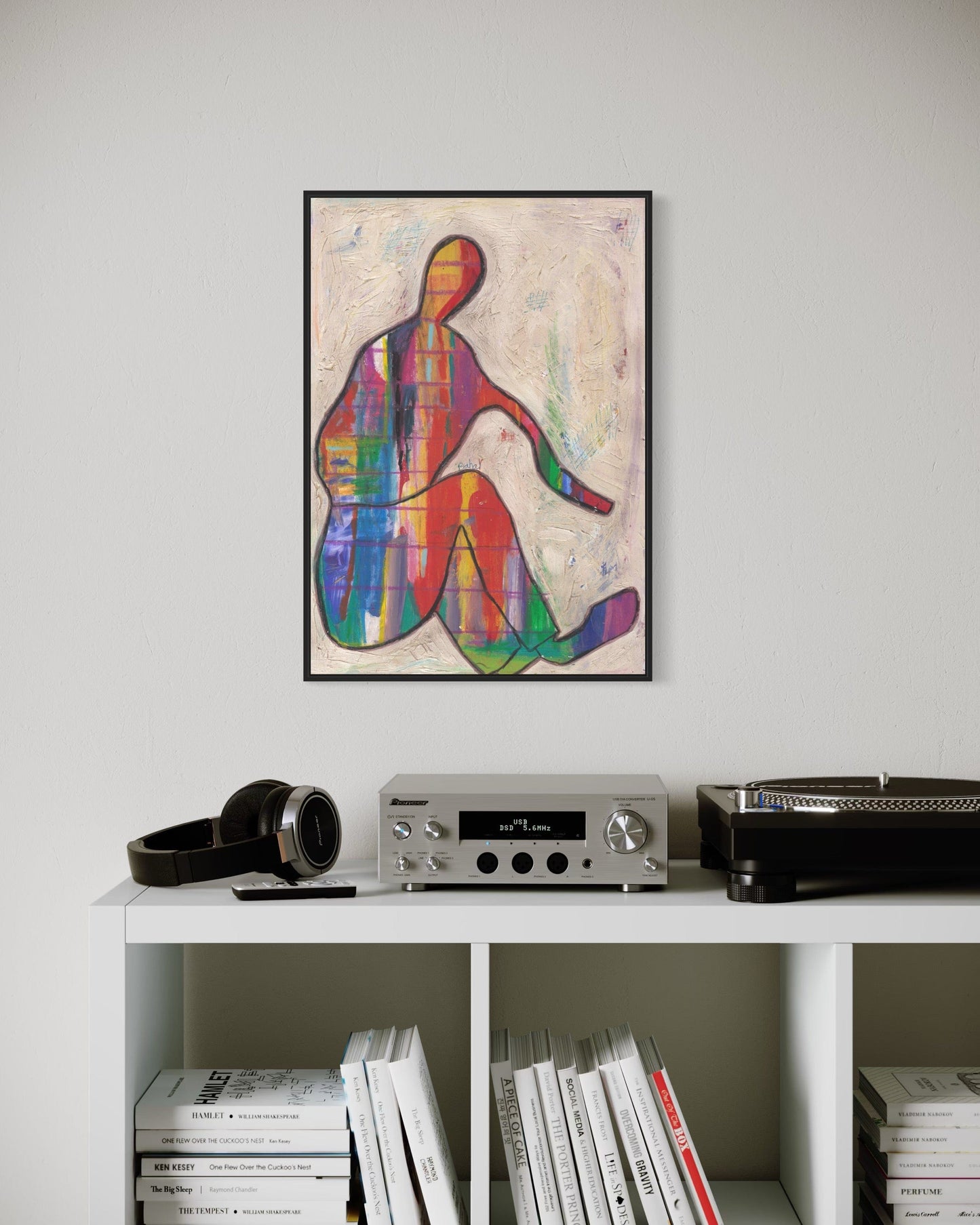 Colorful abstract art print on a wall above a white shelf with books and electronic equipment.
