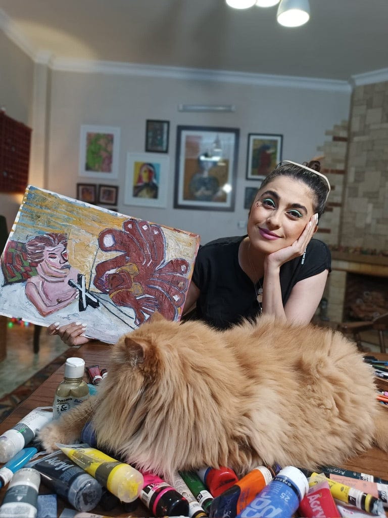 Bahar Haghverdi with a cat surrounded by art supplies in a room with framed pictures on the wall.