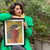 Person holding a framed artwork outdoors with greenery in the background