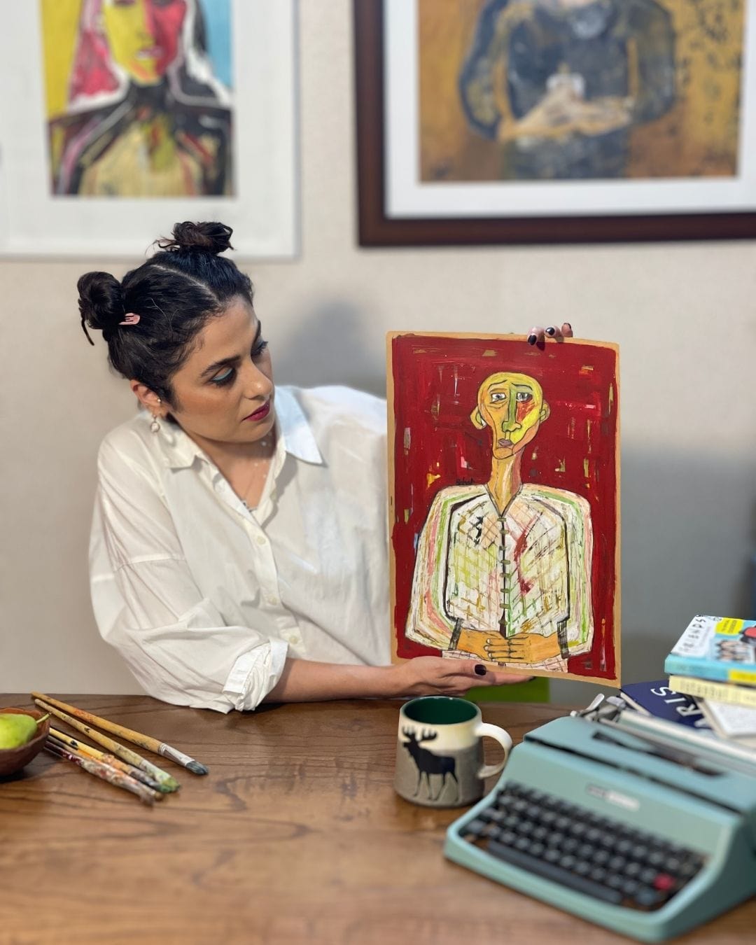 Woman holding a colorful book with an illustration of a person with a yellow head, sitting at a desk with art supplies and a typewriter.