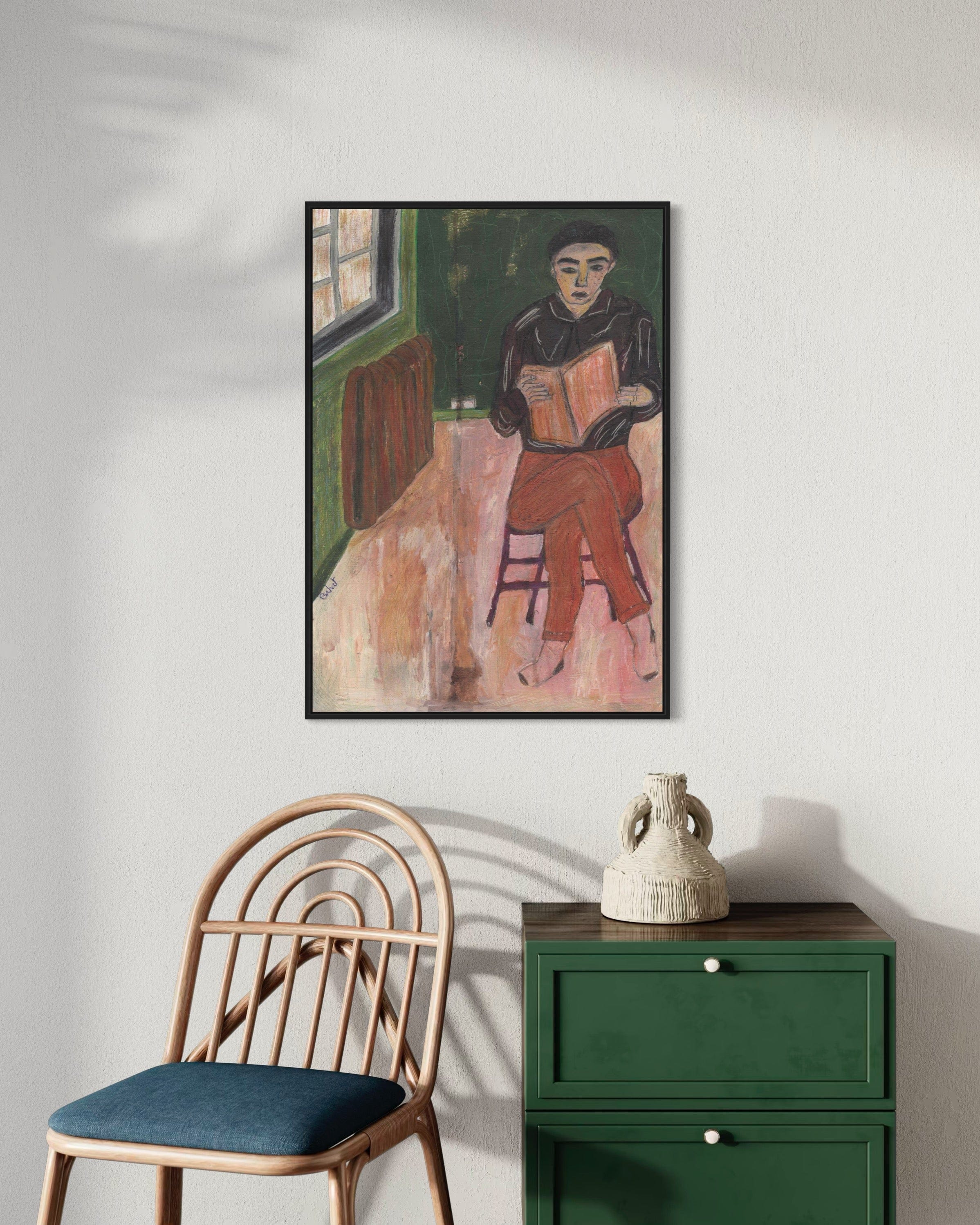 Framed artwork of a person reading a book on a wall above a chair and green cabinet.