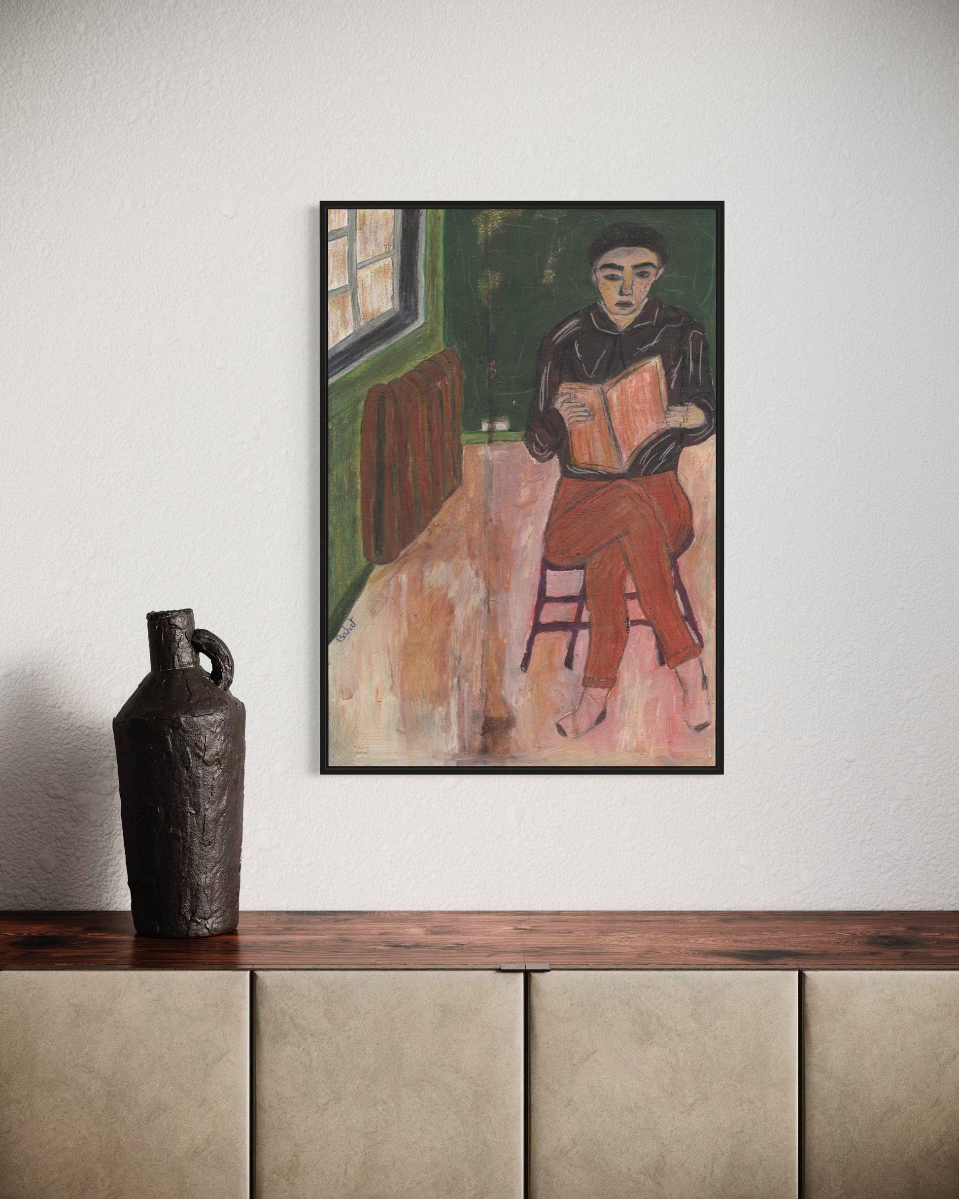 Framed painting of a person reading a book on a wall above a wooden shelf with a vase.