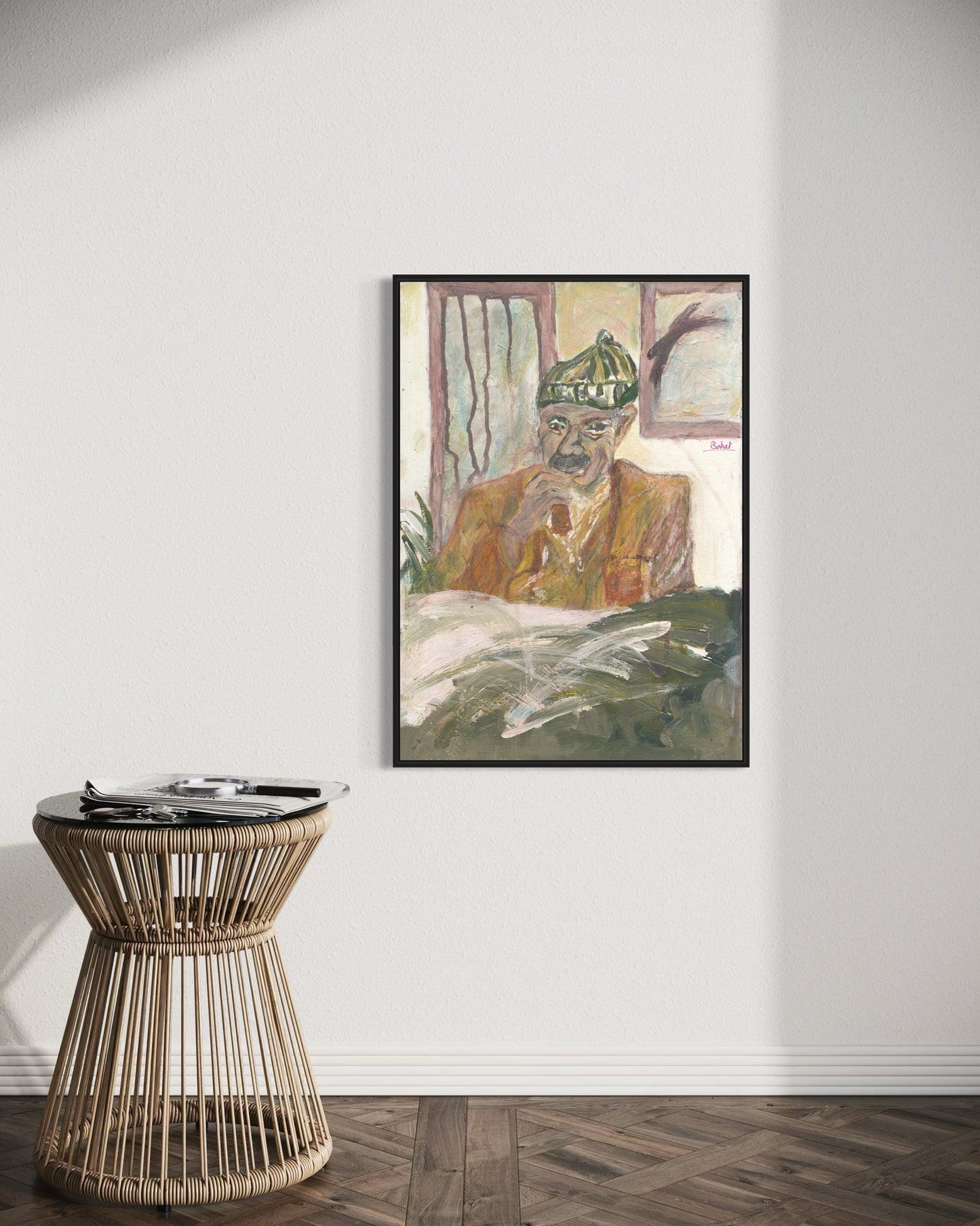 Framed artwork of a person with a green hat on a white wall above a wicker side table.