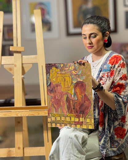Woman holding a painting in an art studio with an easel and framed artwork in the background.